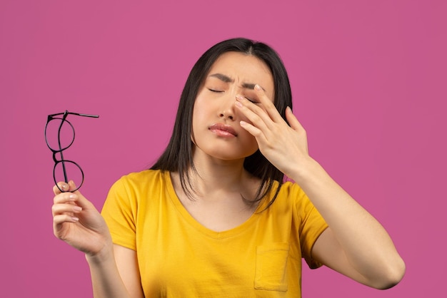 Young asian lady suffering from eye pain taking off glasses and\
touching her eye standing over