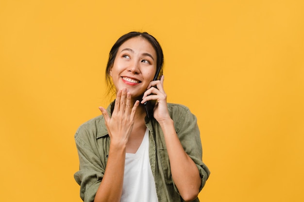 Young Asian lady talk by phone with positive expression, smile broadly, dressed in casual clothing feeling happiness and stand isolated on yellow wall. Happy adorable glad woman rejoices success.