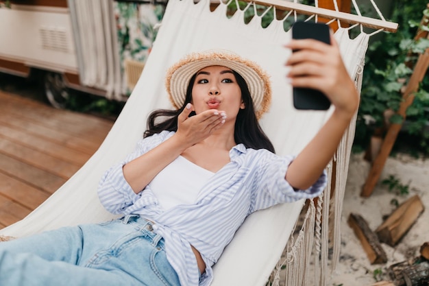 Giovane donna asiatica che chatta in video sullo smartphone inviando un bacio d'aria alla fotocamera mentre giaceva sull'amaca vicino