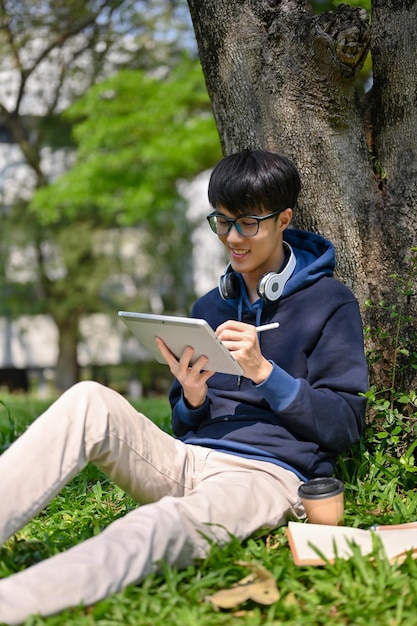 Young Asian male college student using his digital tablet under the tree in the campus park