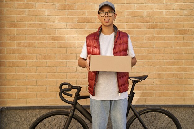 Young asian male courier holding parcel and looking at camera wh