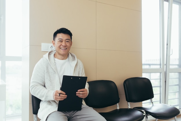 Young asian male in waiting room for interview or meeting holding paper while sitting at chair fills out a resume questionnaire in office