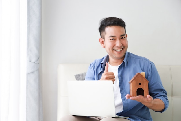 Young Asian Man in blue shirt with Laptop computer and little house model showing for bank loan for house concept in living room