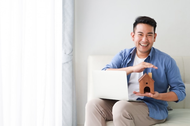 Молодой азиатский человек в голубой рубашке с показом модели портативного компьютера и меньшего дома для банковской ссуды для концепции дома в гостиной