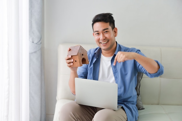 Young Asian Man in blue shirt with Laptop computer and little house model showing for bank loan for house concept in living room