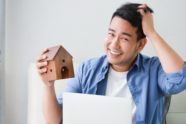 Young Asian Man in blue shirt with Laptop computer and little house model showing for bank loan for house concept in living room