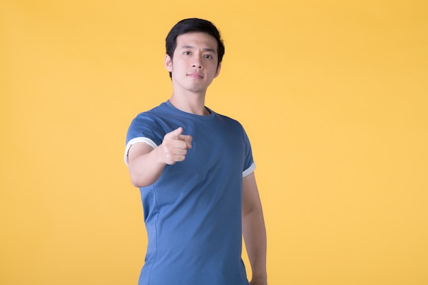 Young Asian man in casual clothes points finger to you and the camera with a confident expression isolated on yellow background