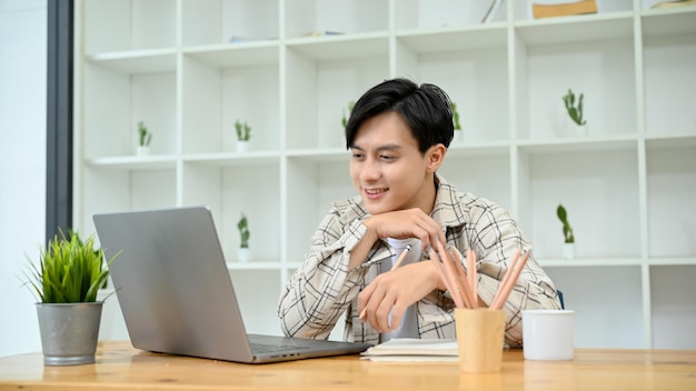 Young Asian man in casual outfit working at his desk looking at laptop screen