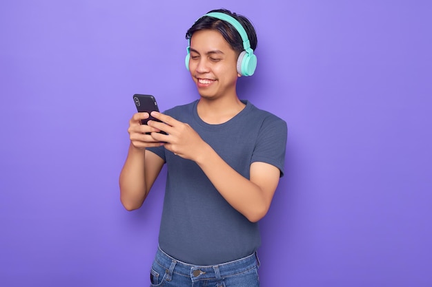 Young asian man in a casual tshirt wearing headphones for listening to music and holding mobile phone isolated over purple background