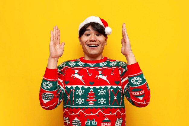 Photo young asian man in christmas sweater and santa hat holds empty hands in front of him