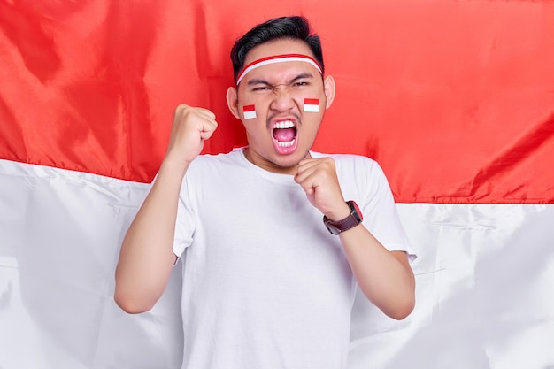 Young Asian man clenched both fist to celebrate Indonesia independence day on August 17 looking at camera with confident and excited expression isolated on indonesian flag background