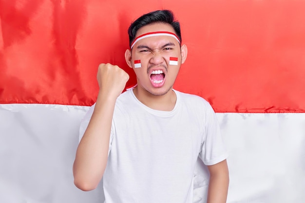 Young Asian man clenched both fist to celebrate Indonesia independence day on August 17 looking at camera with confident and excited expression isolated on indonesian flag background