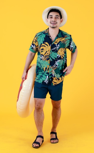 Young Asian man in colorful printed Hawaiian shirt smiles with happiness while holding swim ring. Full body studio portrait on yellow background. Summer holiday travel concept.