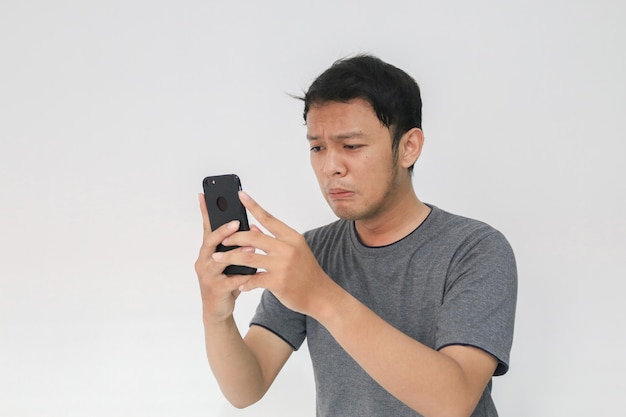 젊은 아시아 남자는 스마트폰을 볼 때 울고 슬퍼합니다. 인도네시아 남자는 검은 셔츠를 입고 격리된 회색 배경