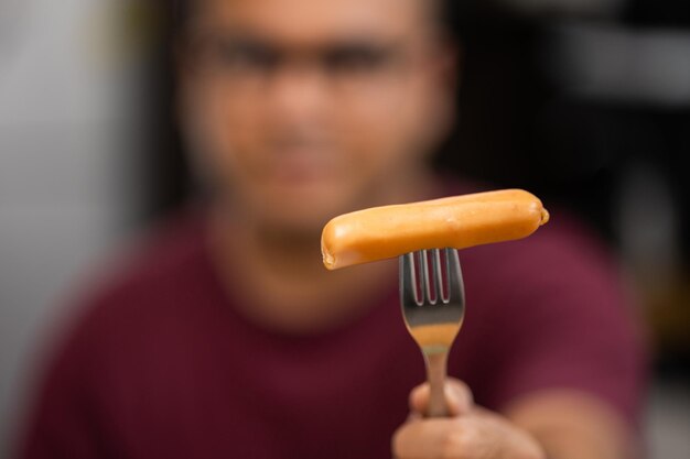 Photo a young asian man eating breakfast, he eats pork sausage. he poked it with a fork and bit his gut and chewed deliciously.