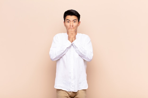 Young asian man feeling worried, upset and scared, covering mouth with hands, looking anxious and having messed up over color wall