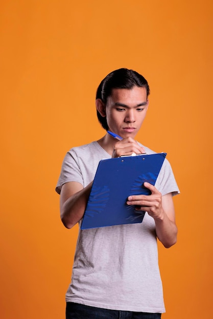 クリップボードに調査票を記入する若いアジア人男性、真面目な学生がメモ帳に書き、メモを取る。集中管理者が書類を保持し、秘書がオフィス プランのチェックリストを作成