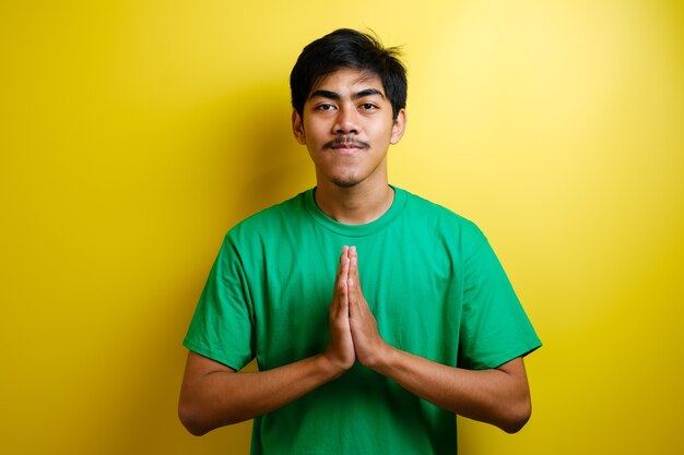 Young Asian Man in green t-shirt smiling and Shows Asian Greeting Gesture against yellow background