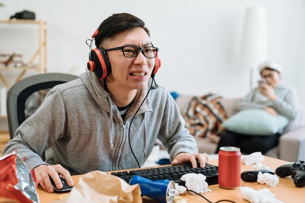 young asian man in headphones addicted to computer online games. concentrated chinese guy typing on keyboard sitting on messy desk. blurred view of girl friend relax using cellphone on sofa in back.