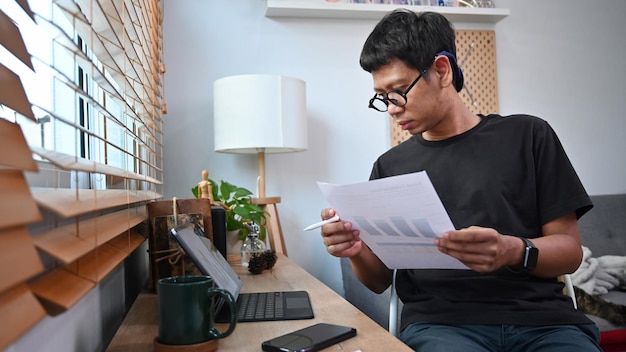Молодой азиатский мужчина держит финансовые графики и статистические документы и работает с компьютерным планшетом дома