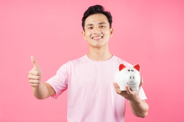 Young Asian man holding piggy bank on pink