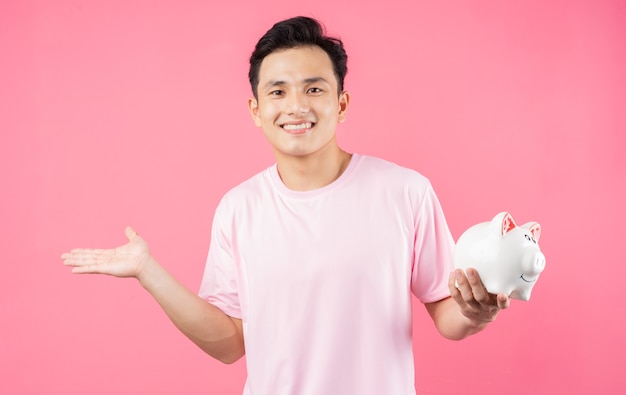 Young Asian man holding piggy bank on pink