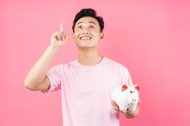 Young Asian man holding piggy bank on pink