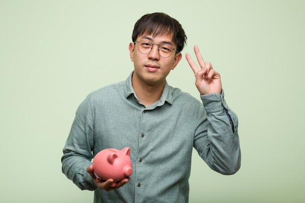 Young asian man holding a piggy bank showing number two