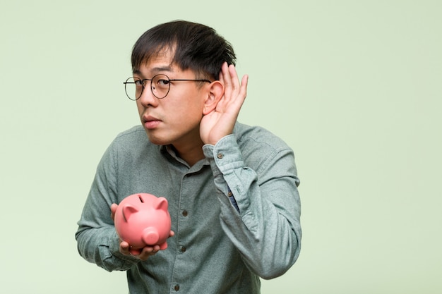 Young asian man holding a piggy bank try to listening a gossip