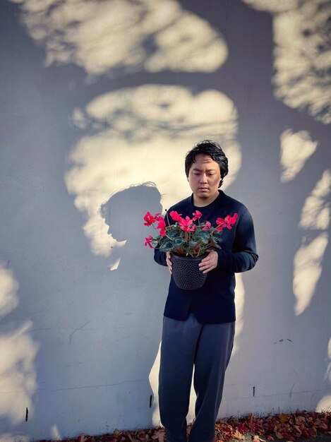 Photo young asian man holding red flowering cyclamen potted plant against shadow patterned wall