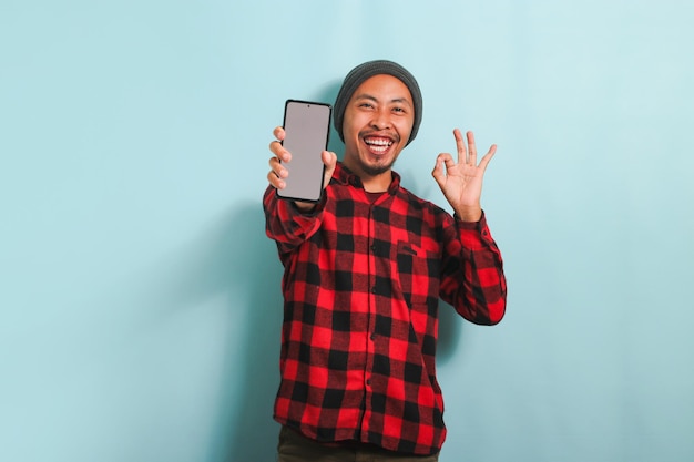 Young Asian man holding smartphone mockup blank screen and shows OK sign isolated on blue background