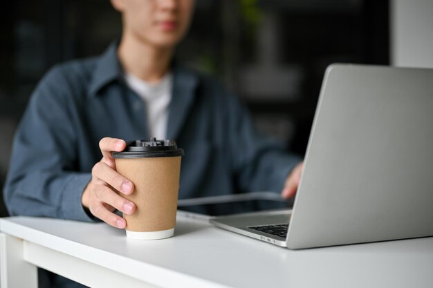 A young Asian man holding a takeaway coffee cup working on his tasks on laptop in coffee shop