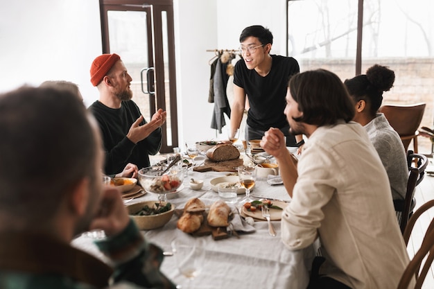 사진 안경과 검은 색 티셔츠에 젊은 아시아 남자가 동료들과 행복하게 이야기하는 테이블에 기대어