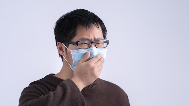 Young Asian man is coughing, feeling unwell, vomit sick with wearing a medical blue face mask isolated on white background, close up, copy space.