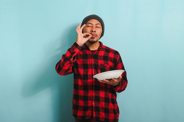 Young Asian man is making delicious hand gesture while holding plate isolated on blue background