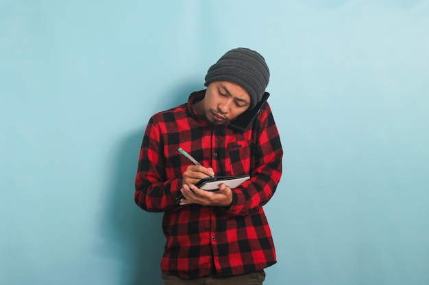 Young Asian man is making notes while talking on his mobile phone isolated on a blue background