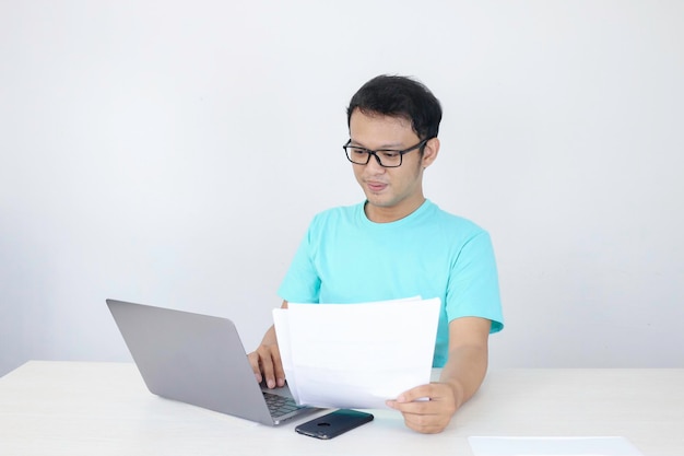 Молодой азиатский мужчина улыбается и счастлив, работая на ноутбуке и документируя под рукой Индонезийский мужчина в синей рубашке
