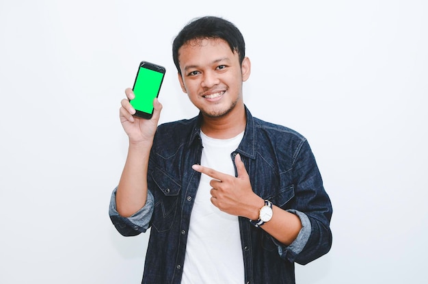 Young Asian man is standing and smiling pointing on green screen on smartphone screen on white background