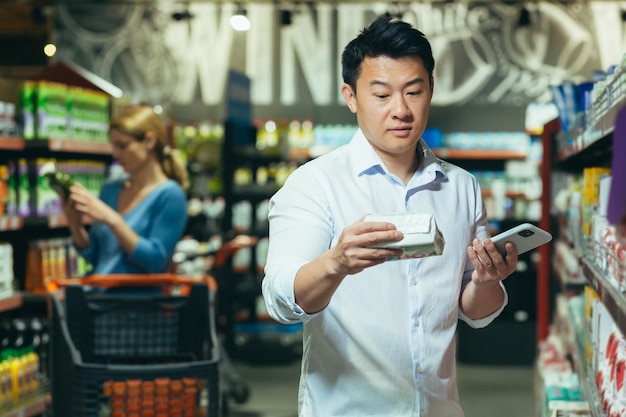 한 젊은 아시아 남성이 슈퍼마켓에 서서 그렇지 않은 아내 여자친구를 위해 패드를 고르고 있다
