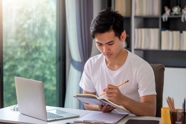 Young Asian man is taking notes on the laptop table at home.