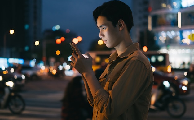 Young Asian man is using his phone while walking in the street at night