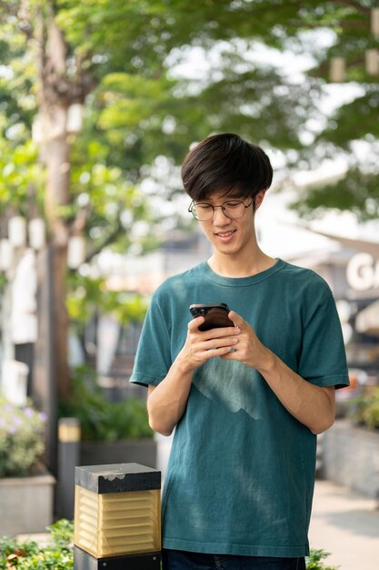 アジアの若い男が公園で友達を待っている間スマートフォンを使って友達とチャットしています