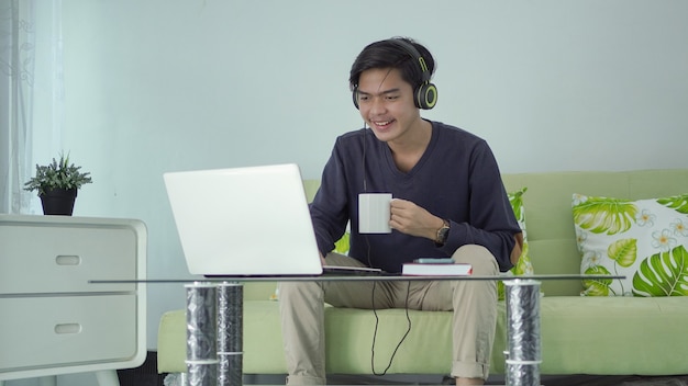 Photo young asian man listening to sound from his laptop with a cup of drink at home