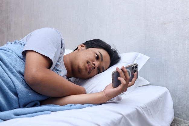 Young Asian man looking at mobile phone on bed before sleeping