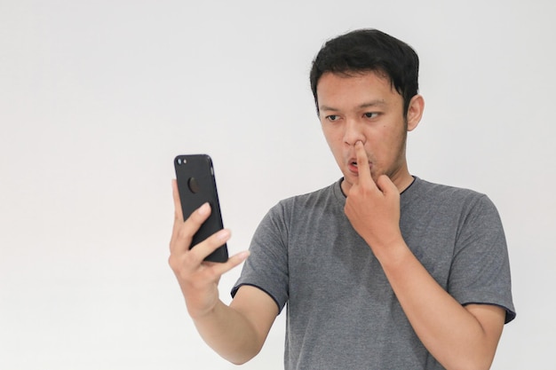 Молодой азиатский мужчина смотрит в смартфон с жестом ковыряния в носу