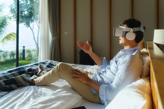 Young Asian man lying on bed and using virtual reality glasses for business meeting during his travel summer vacation holiday