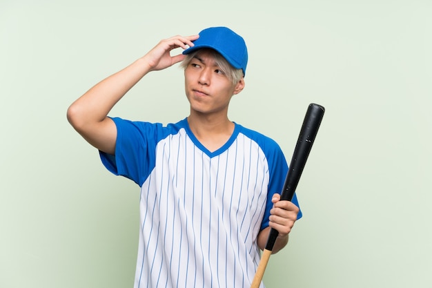 Молодой азиатский человек играя бейсбол над изолированным зеленым цветом имея сомнения и с смущает выражение стороны