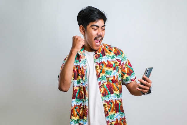 Молодой азиатский человек позирует на изолированном белом фоне, играет в игры по телефону, делает жест победителя.