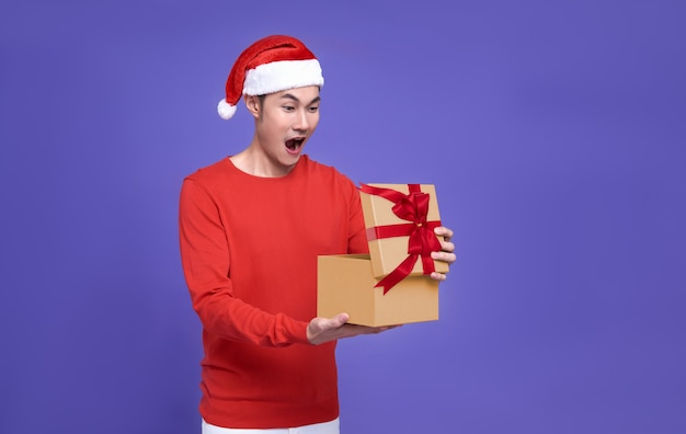 Young asian man in red casual attire wearing Santa hat opening presents box and looking excited and surprise something inside box isolated on purple wall.Happy new year concept.