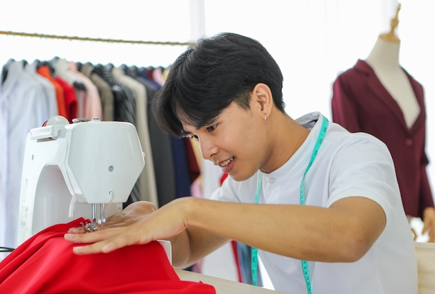 現代のプロのアトリエで働いている間、機械で赤いアパレルを縫う若いアジア人男性。